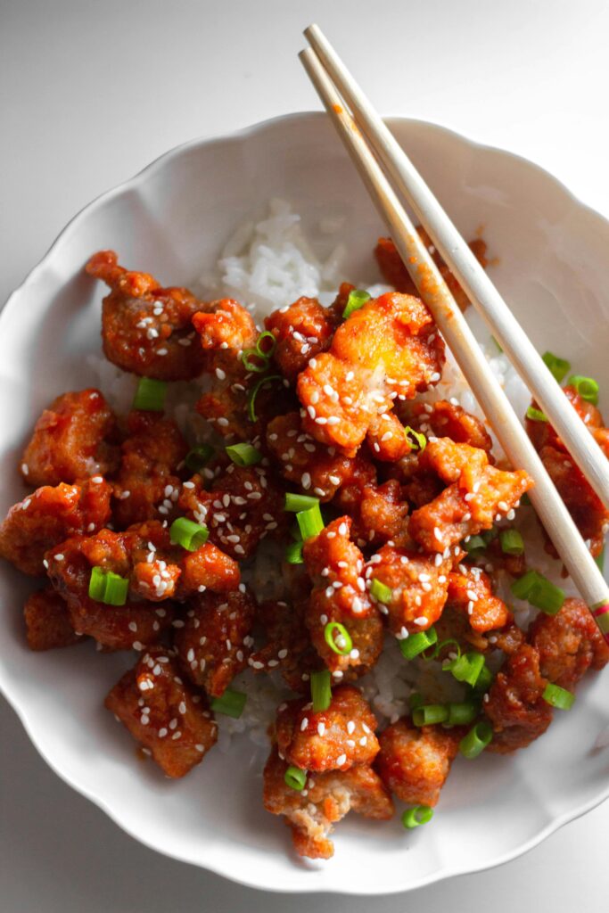 Crispy chicken pieces with sesame and scallions over rice, served with chopsticks.